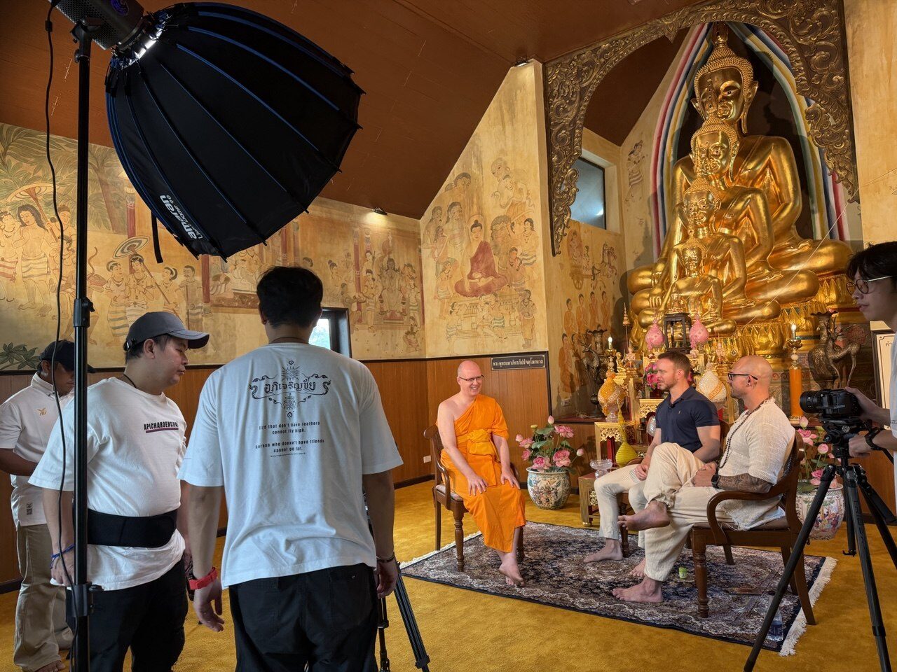 A film crew captures a monk of iMonastery seated before a golden Buddha inside a temple hall at the Ancient City near Bangkok